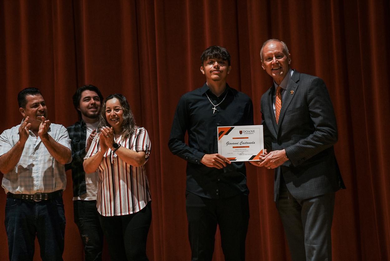 Giovanni Castañeda holds his scholarship announcement next to Dr. Roger Hughes, president of Doane University. Castañeda's family stands back to the left, clapping.