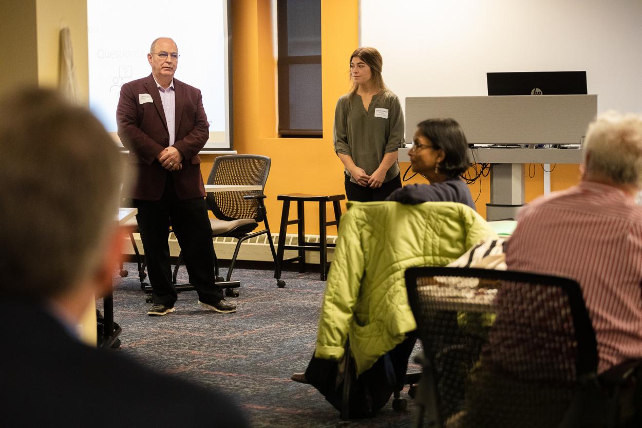 Pete Poppert, assistant professor of practice in agribusiness and Sydney Erickson, junior business administration major, stand at the front of a yellow-painted classroom to answer questions from guests.