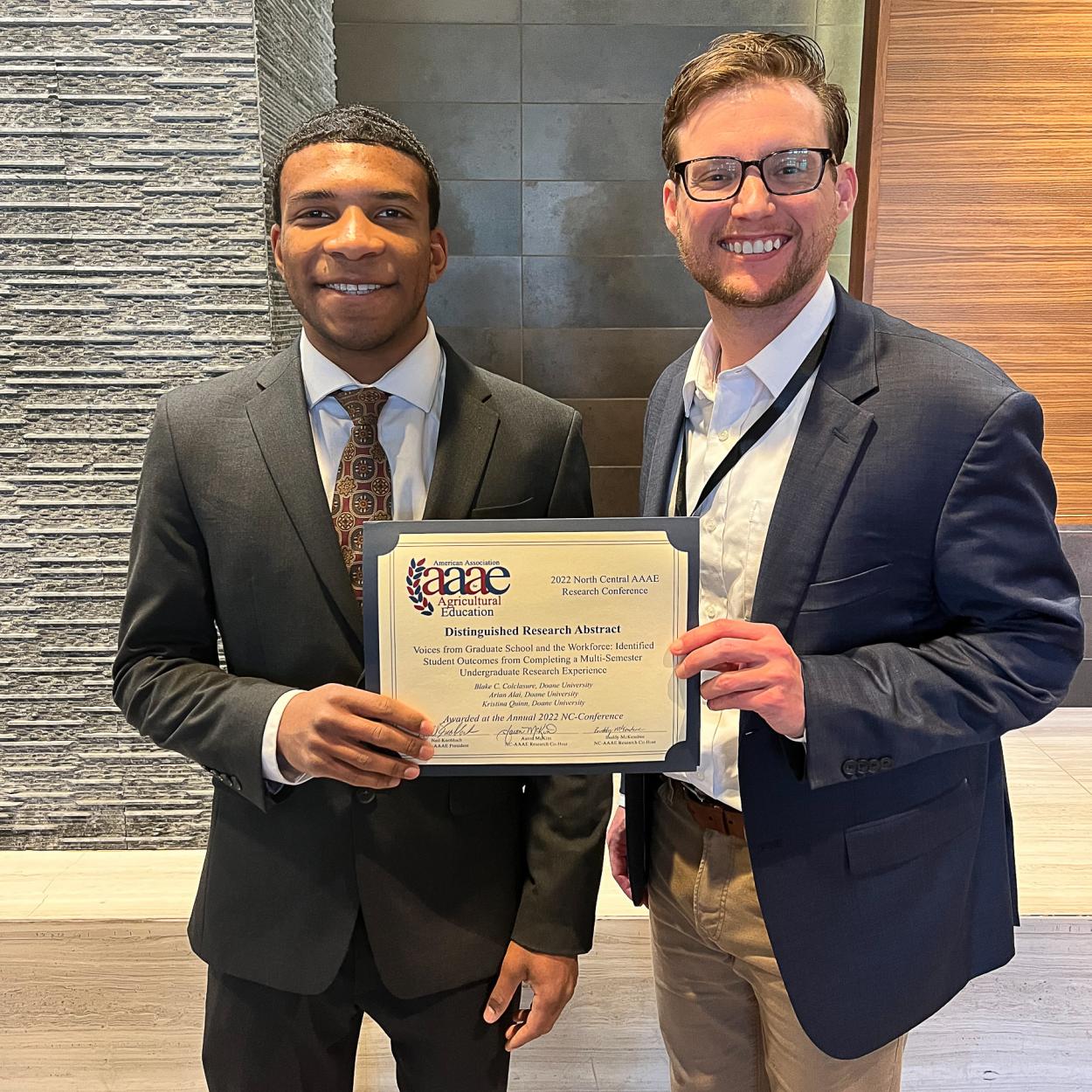 Arian Alai ’23 stands in a grey suit, white shirt and red and yellow tie next to Dr. Blake Colclasure, who wears a blue jacket over a white shirt and khaki pants. They hold an award that reads "Distinguished Research Abstract."