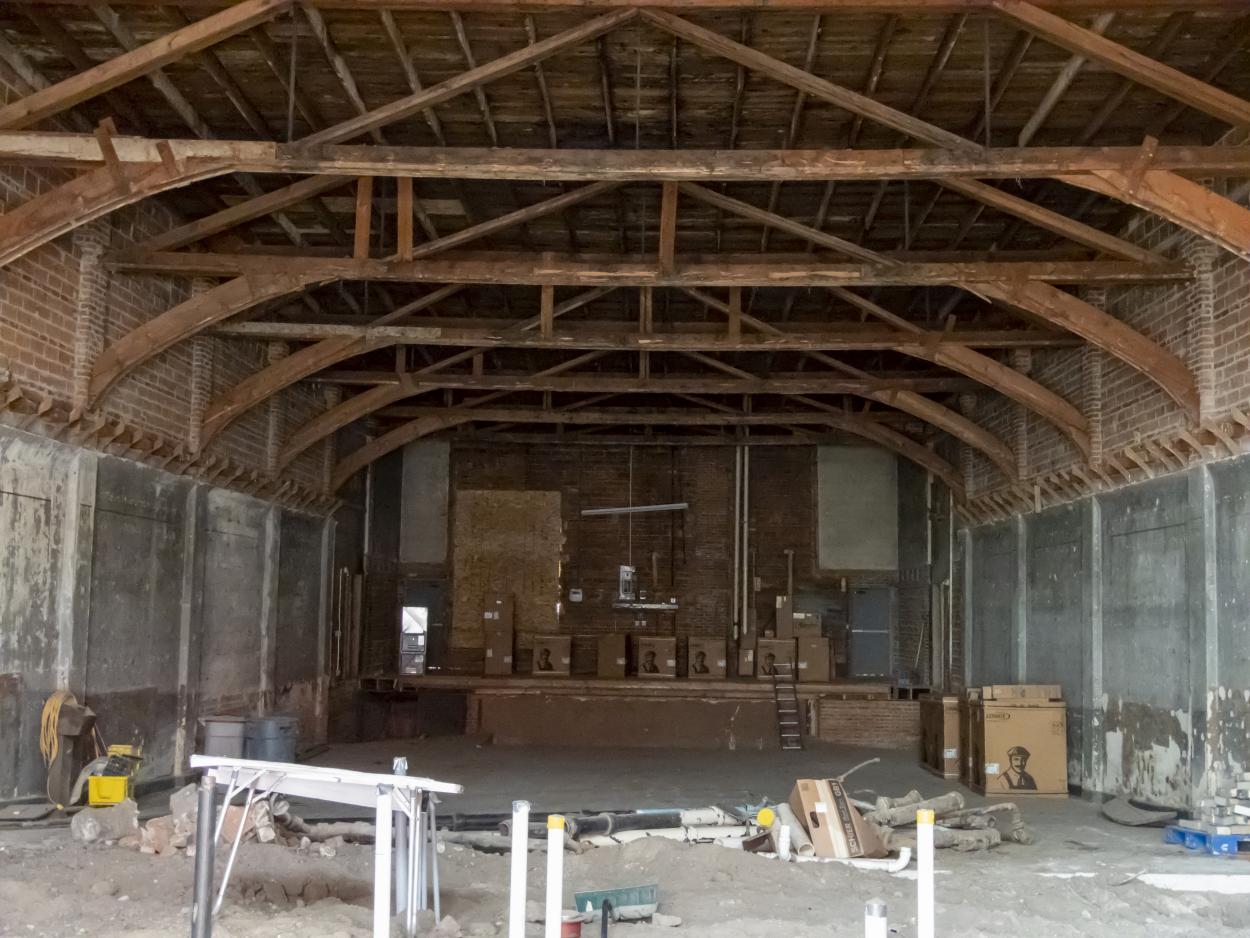 Image of the interior of the Isis Theatre in Crete, Nebraska, which is currently undergoing renovation. The arched ceiling beams are exposed and walls are down to studs and brickwork, but it's very clearly still a theatre, with a stage at the far end of the building. 