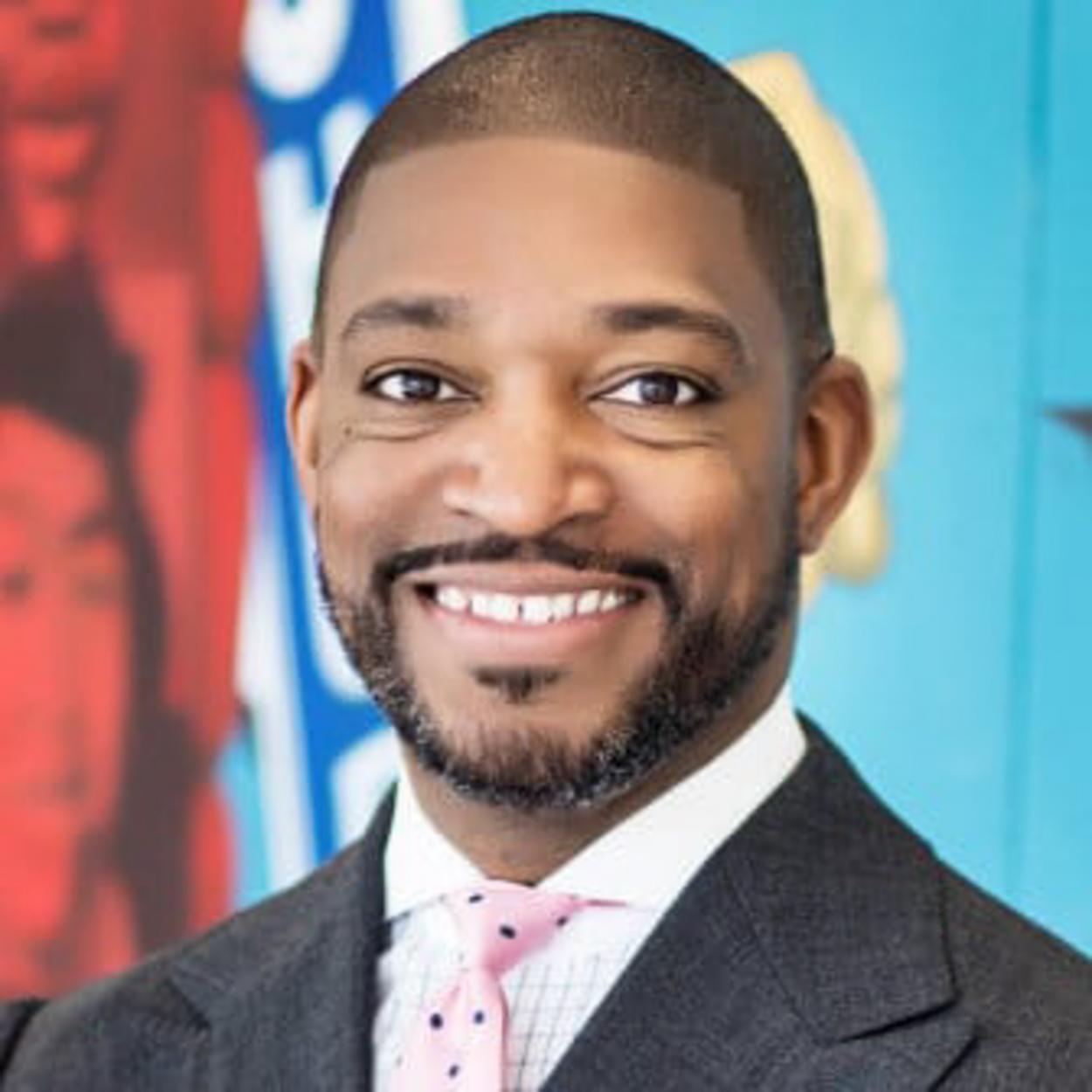 Portrait of Rev. Dr. Starsky Wilson, a Black man smiling broadly in front of a colorful background. He has closely trimmed hair, and a closely trimmed beard, and is wearing a dark grey suit, a white shirt, and a light pink tie with small black dots. 