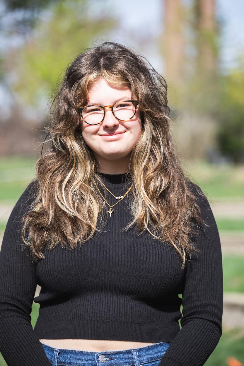 Abrianna Miller stands in front of Cassel Theatre, a grassy, terraced amphitheatre. Her curly brown hair has highlights and reaches past her shoulders. She wears round tortoiseshell glasses, a black, cropped mockneck shirt with two gold necklaces. 
