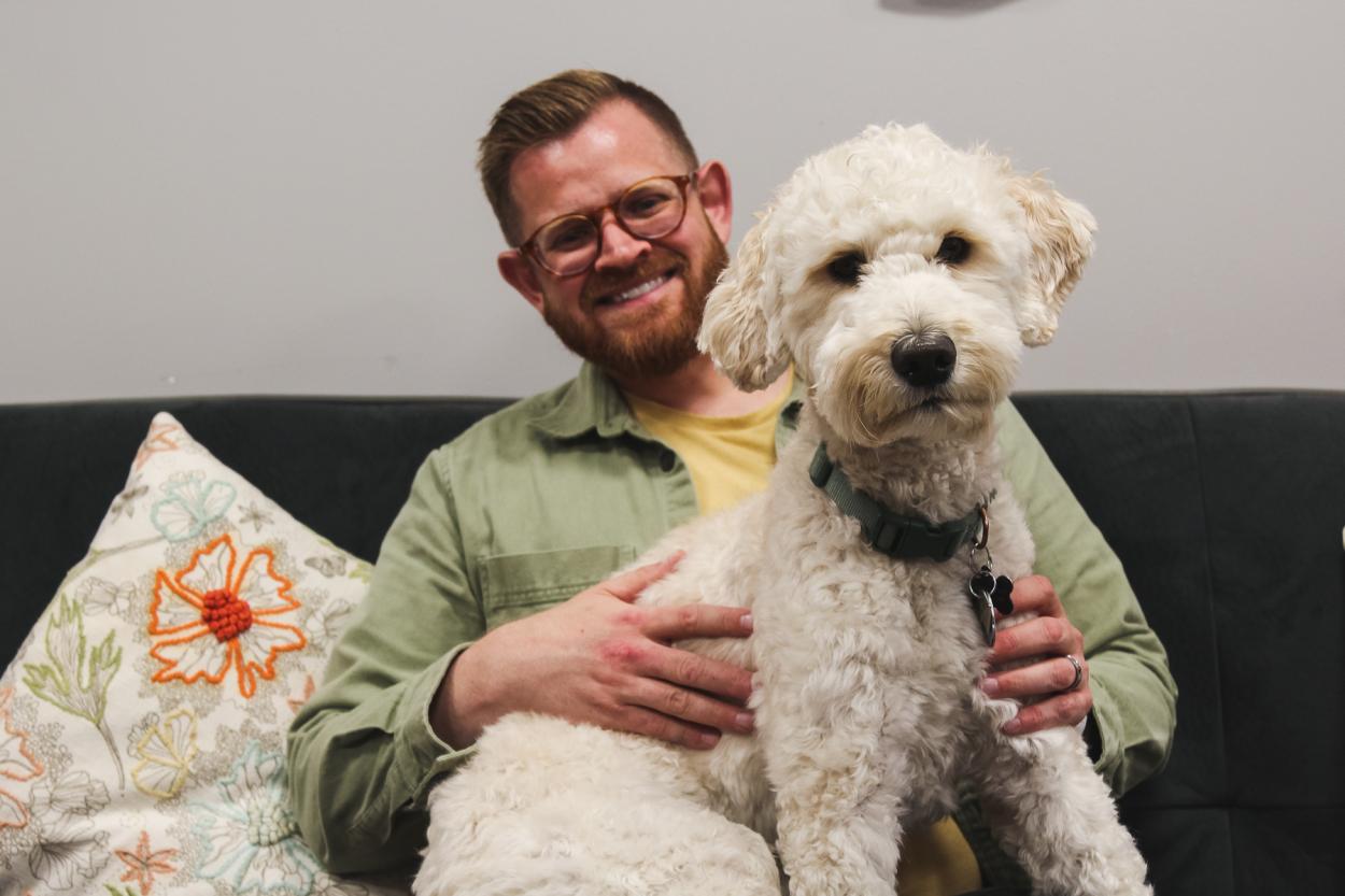 Georgie, a light-colored goldendoodle with fluffy trimmed fur, sits on the lap of his person, Blake Tobey, academic specialist in the Academic Success Center. The two sit on a comfy grey couch in the center's office in the Communications Building. 