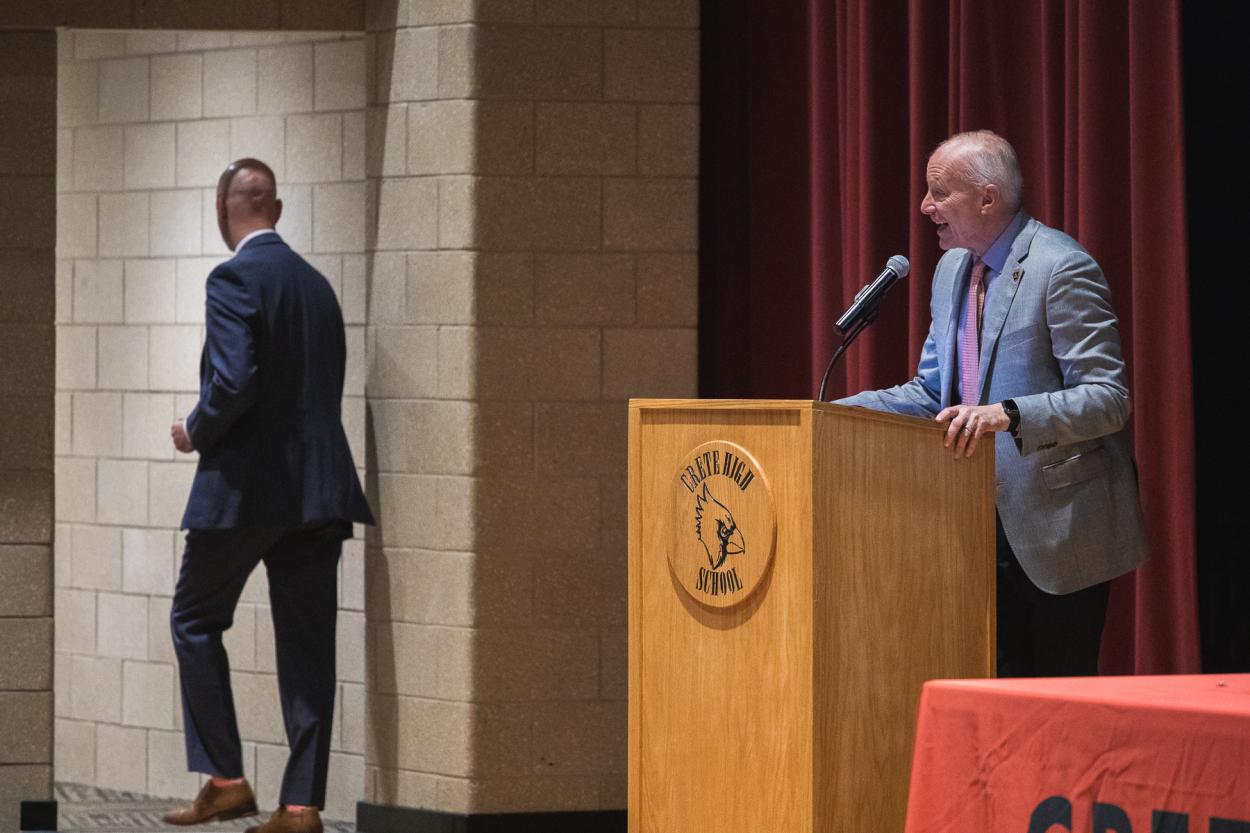 Doane President Dr. Roger Hughes addresses employees of Crete Public Schools behind a wooden podium with the Crete High School cardinal head logo. CPS Superintendent Dr. Joshua McDowell walks offstage through a doorway. Behind Hughes is a red curtain. 