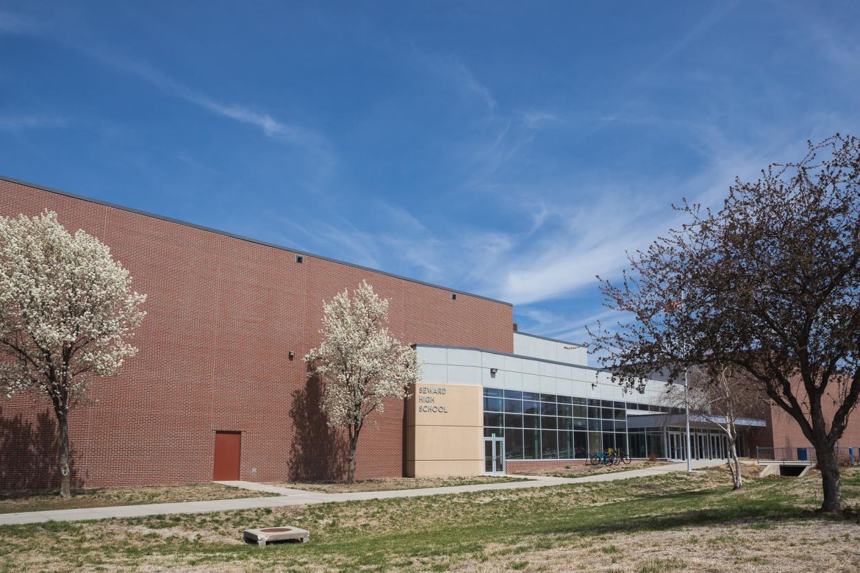 The exterior of Seward High School, a red brick building with an entrance made of glass windows and doors, and grey and stone-colored blocks. The grass is just turning green, two trees are blooming with white flowers and the sky overhead is bright blue with wispy clouds.