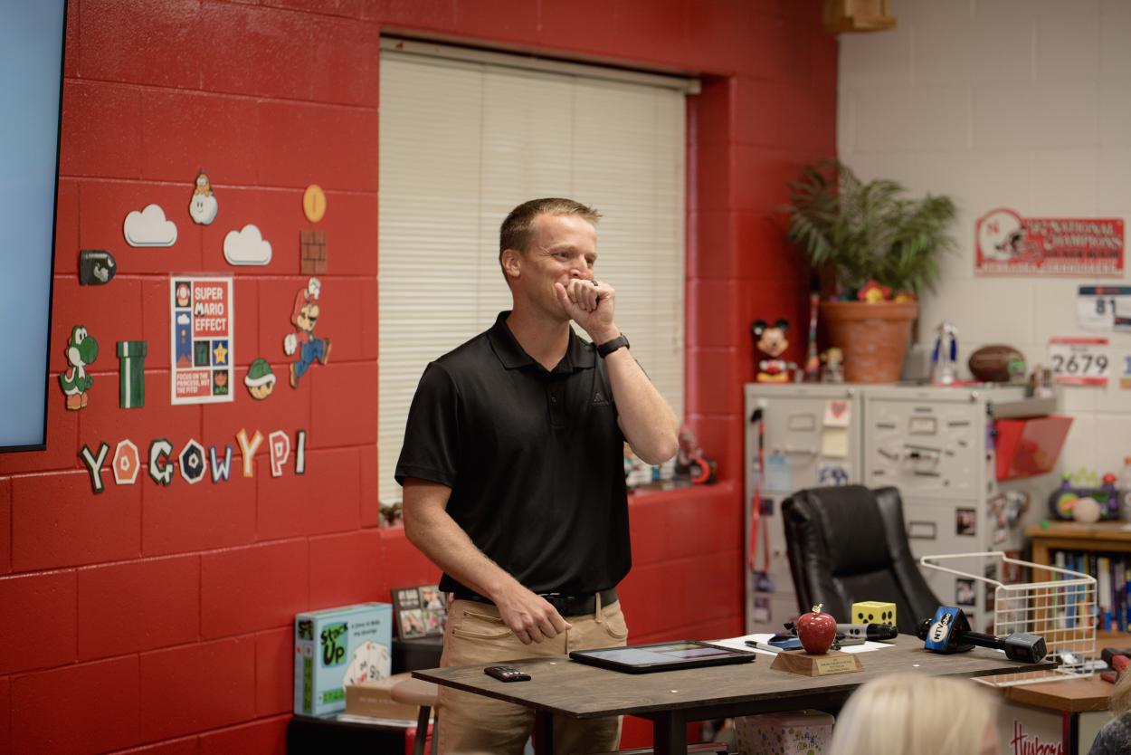 Aurora Middle School teacher Scott Phillips, ’16E, reacts to learning he is the 2024 Nebraska Teacher of the Year after a surprise presentation during one of his seventh-grade math classes. Phillips stands in front of a window with blinds closed, behind a desk in a classroom with red-painted walls. He wears a black polo tshirt and has his hand up to his mouth in an expression of surprise. 