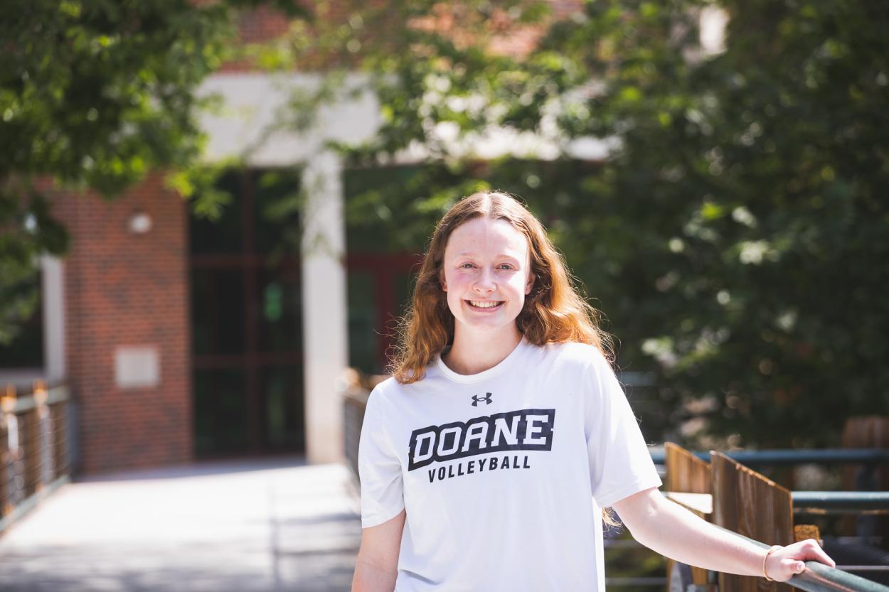 Junior MacKenzie Painter wears a white tshirt that reads "Doane Volleyball." She stands in front of the Lied Science and Mathematics Building.