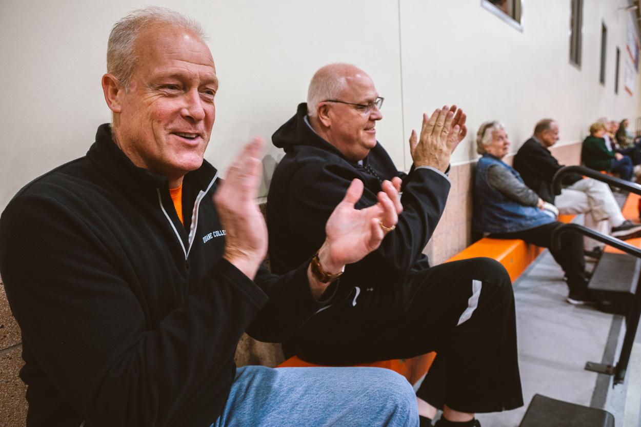 Tom Sorensen (left) claps for the Doane Women's Basketball team. He wears a black quarterzip with a "Doane College" logo, and sits at the top of the orange bleachers next to Marty Fye.