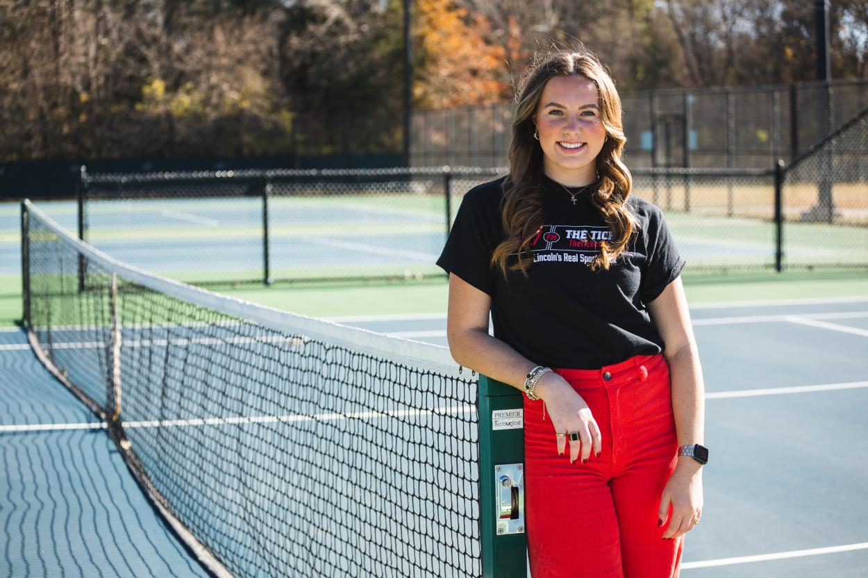 Image of Grace Schroller, leaning against a green pole for the tennis nets on Doane's blue tennis courts. Schroller wears a black shirt that reads "93.7 The Ticket" and red corduroy pants.