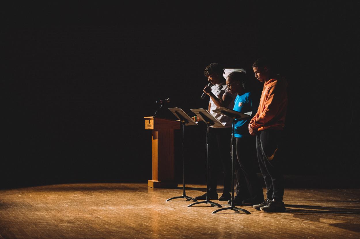 Three Black students, wearing casual clothing, stand in a row behind black music stands to read from Rev. Dr. Martin Luther King Jr.'s 1963 "Letter from Birmingham Jail." The students are lit by a spotlight, with all but the wood stage in front of them and a wooden podium to their right, in shadow. They are participating in a candlelight vigil, one of the events of Doane's MLK Week.