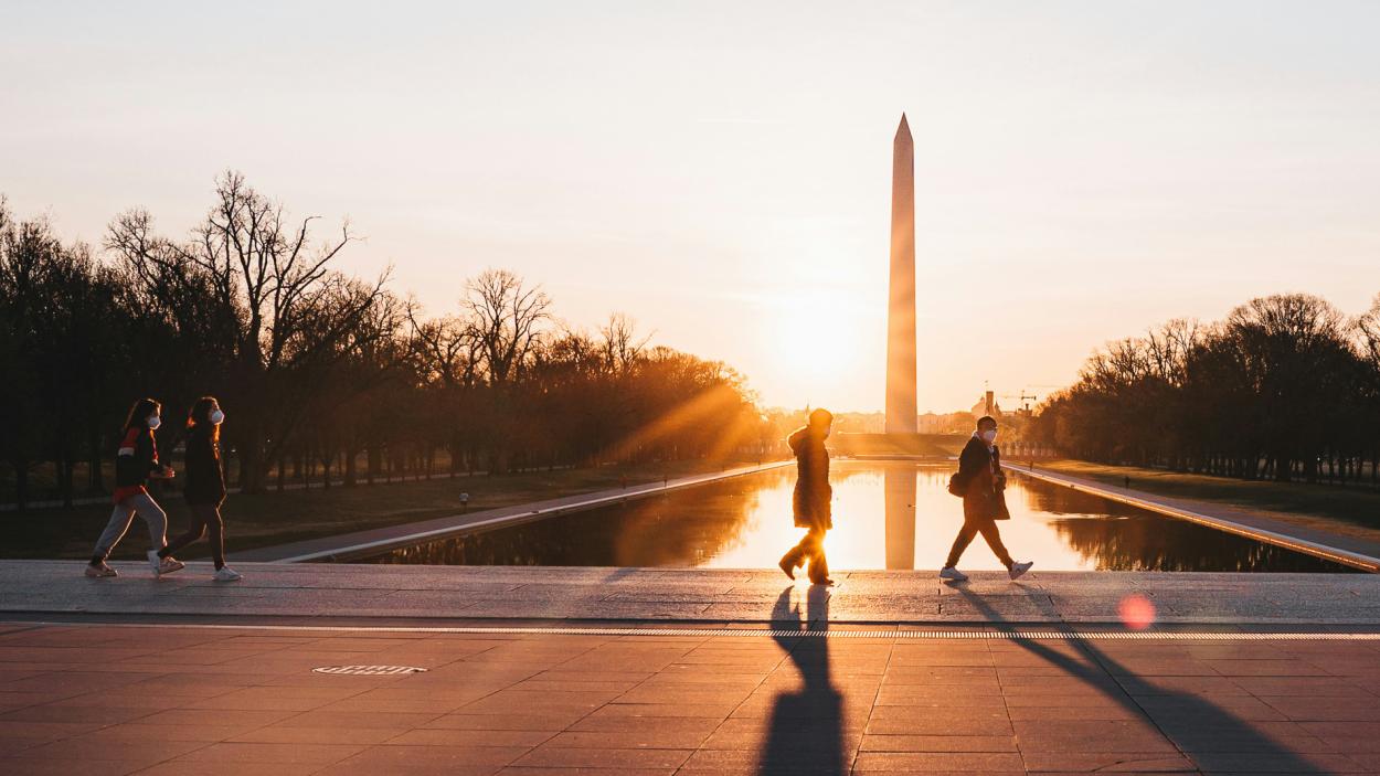 Image by Eric Dekker for Unsplash. Four people are backlit by the sunrise as they walk in front of a long rectangular pool. At the far edge of the pool stands a tall white obelisk, and trees grow to the left and right of the pool. 