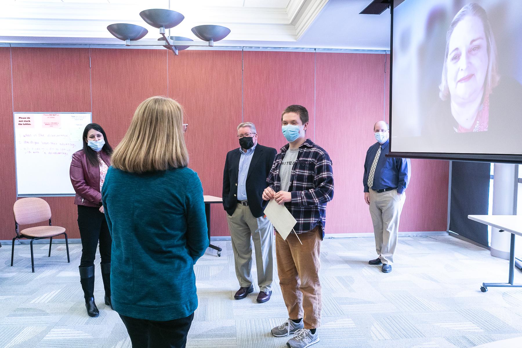 Samuel Province (middle, plaid shirt) speaks with Nancy Murphy, financial aid counselor. He holds a certificate announcing his scholarship. The donor for whom the scholarship is named, Carolyn Kollmeier '74, joined by Zoom and is shown on a screen on the right side of the image.