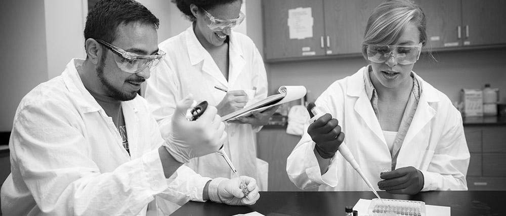 Students titrating a solution in lab coats and safety glasses while an instructor takes notes in the background