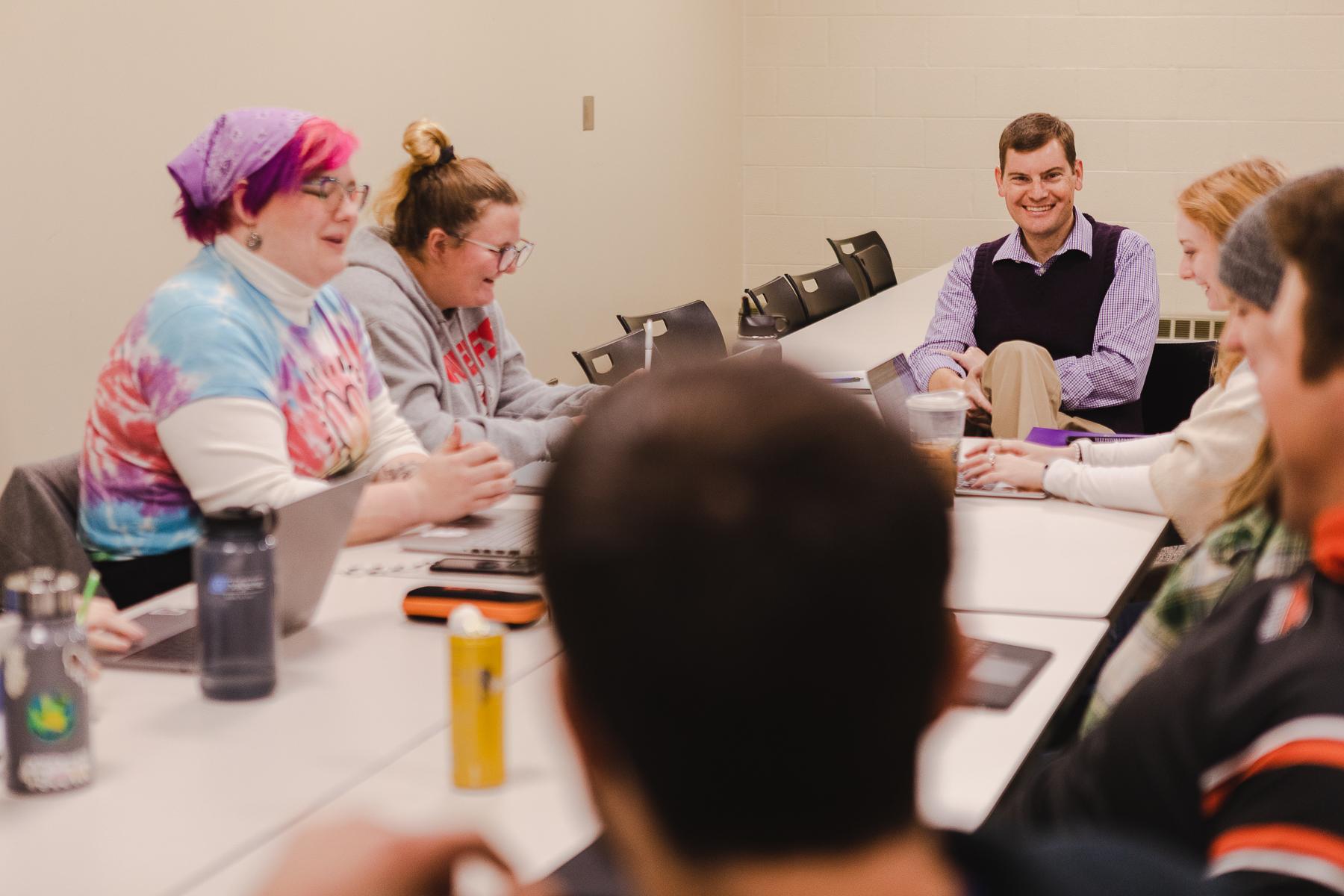 Dr. Timothy Hill, professor of political science, sits at the end of a long table with laughing and smiling students during a class.