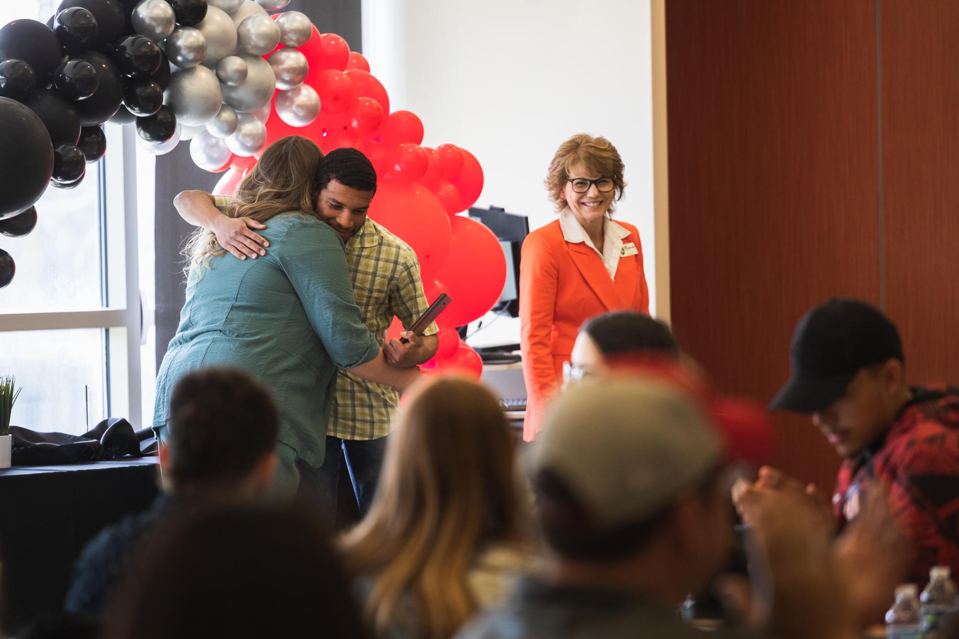 Senior Arian Alai, wearing a green plaid shirt, hugs Jill Kline, wearing a teal jacket, in front of a black, silver and red balloon arch. In the foreground, students and their families are seated, with only Arian and Jill in focus.