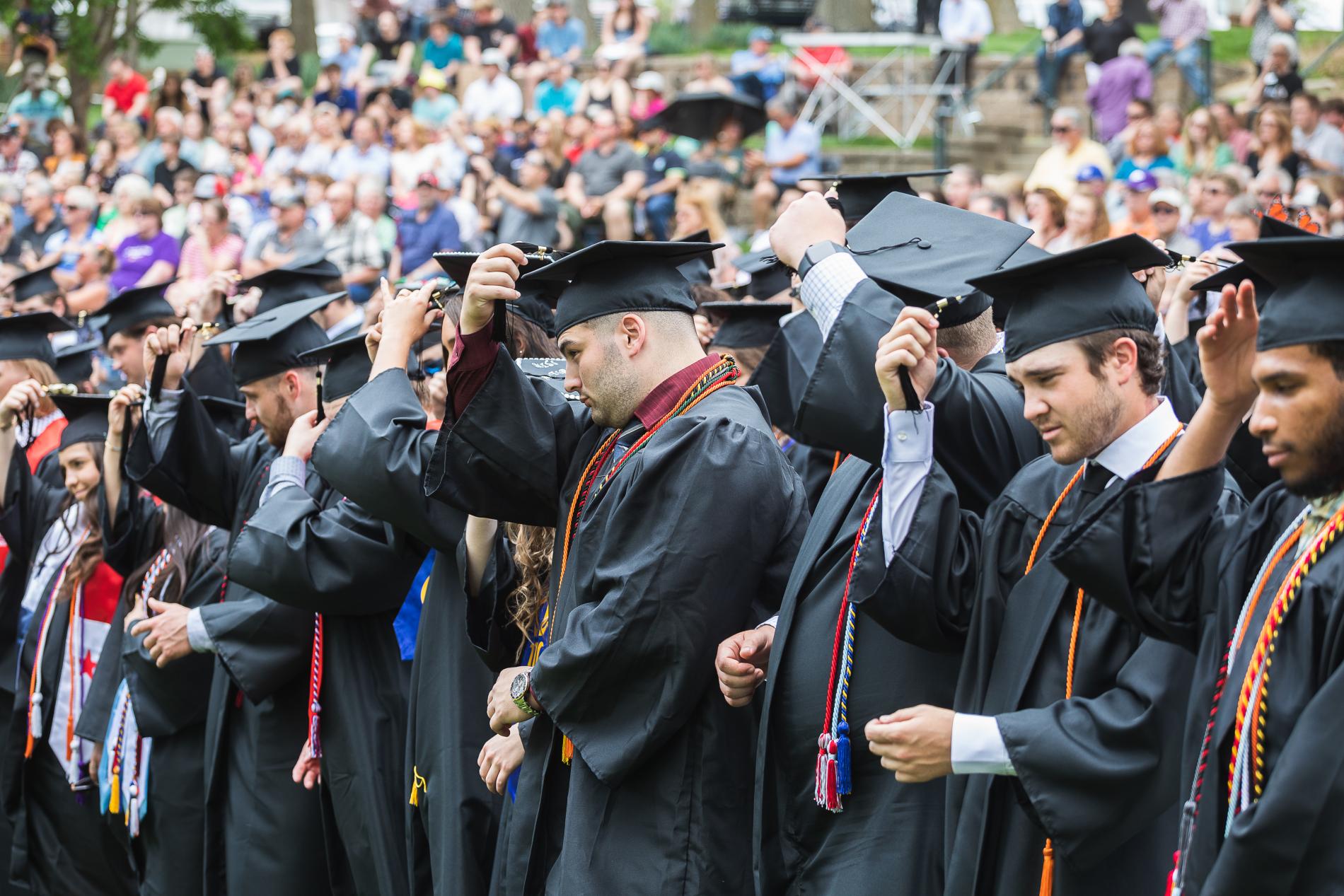 A row of Doane graduates at the spring 2023 afternoon commencement ceremony, clad in black gowns and black caps with bright sashes and cords, turn their tassels to signify receiving their degrees. Blurred in the background are their families and friends seated in Cassel Theatre, a green, grassy amphitheater that rises around the graduates. 