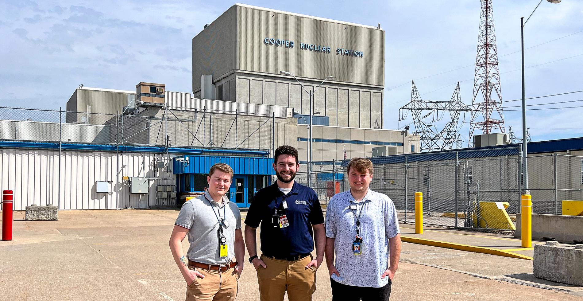 Three men in polos with badges stand on a concrete parking lot in front of a blocky concrete building bearing the words "Cooper Nuclear Station."