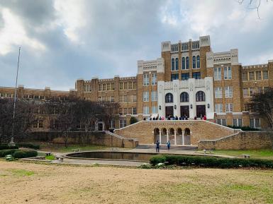 An image of the front of Central High School, in Little Rock, Arkansas. The school is yellow brick with a reflecting pool and grass lawn in front. Tourists mill around the pond and up stairs to the doors of the school.