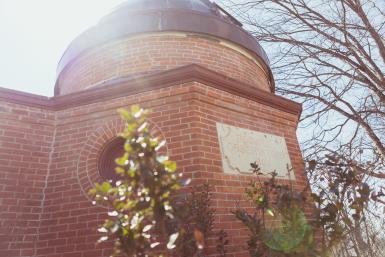 Image of Boswell Observatory, a red brick building topped by a copper dome that opens and closes for skyviewing. A plaque on the building reads "Boswell Observatory 1883." The image is from a low angle, with a few plants in the foreground and the building looming above. 