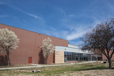 Image of the exterior of Seward High School, a red brick building with an entrance made of glass windows and doors, and light grey and stone-colored blocks. Trees in front are blooming and the grass is turning green. The sky overhead is bright blue with wispy clouds. 