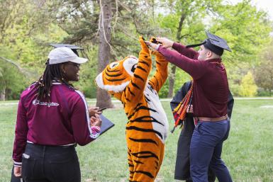 Thomas the Tiger, Doane's mascot, gives a double high-five to Brandon Antesberger after the university's spring 2023 afternoon commencement ceremony. Antesberger still wears his cap and tassel, unknowingly high-fiving a fellow graduate, Trey Porter, who served as Doane's mascot from 2019-2023.