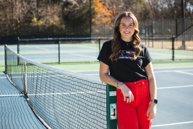 Grace Schroller leans against a green pole holding up a tennis net on Doane's blue-painted courts. She wears a black t-shirt with the logo for "The Ticket" and red corduroy pants.