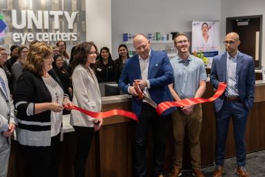 Doctors and staff in business casual clothing at Unity Eye Centers hold a red ribbon for Chad Hudnall O.D., wearing a blue suit with a white shirt, who cuts the ribbon celebrating Unity and Allen Capital Group's grand opening in Grand Island. In the background is a sign for Unity Eye Centers. The walls are painted white.