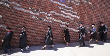 Seven students in black caps and gowns walk down a ramp in front of a brick wall showing a mural made of wavy tile.